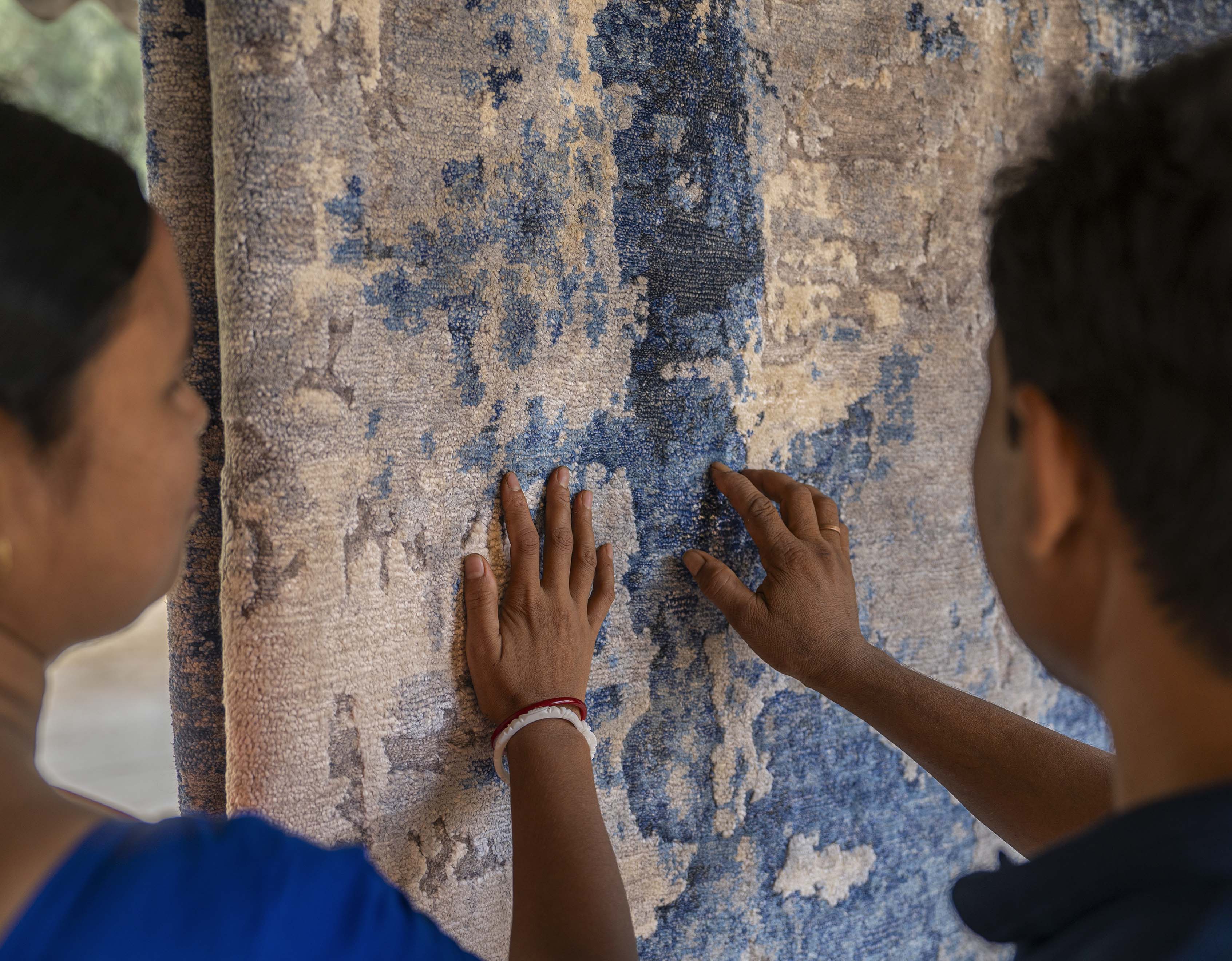 Two people touch a textured, patterned blue and beige rug with both hands, examining its surface closely.