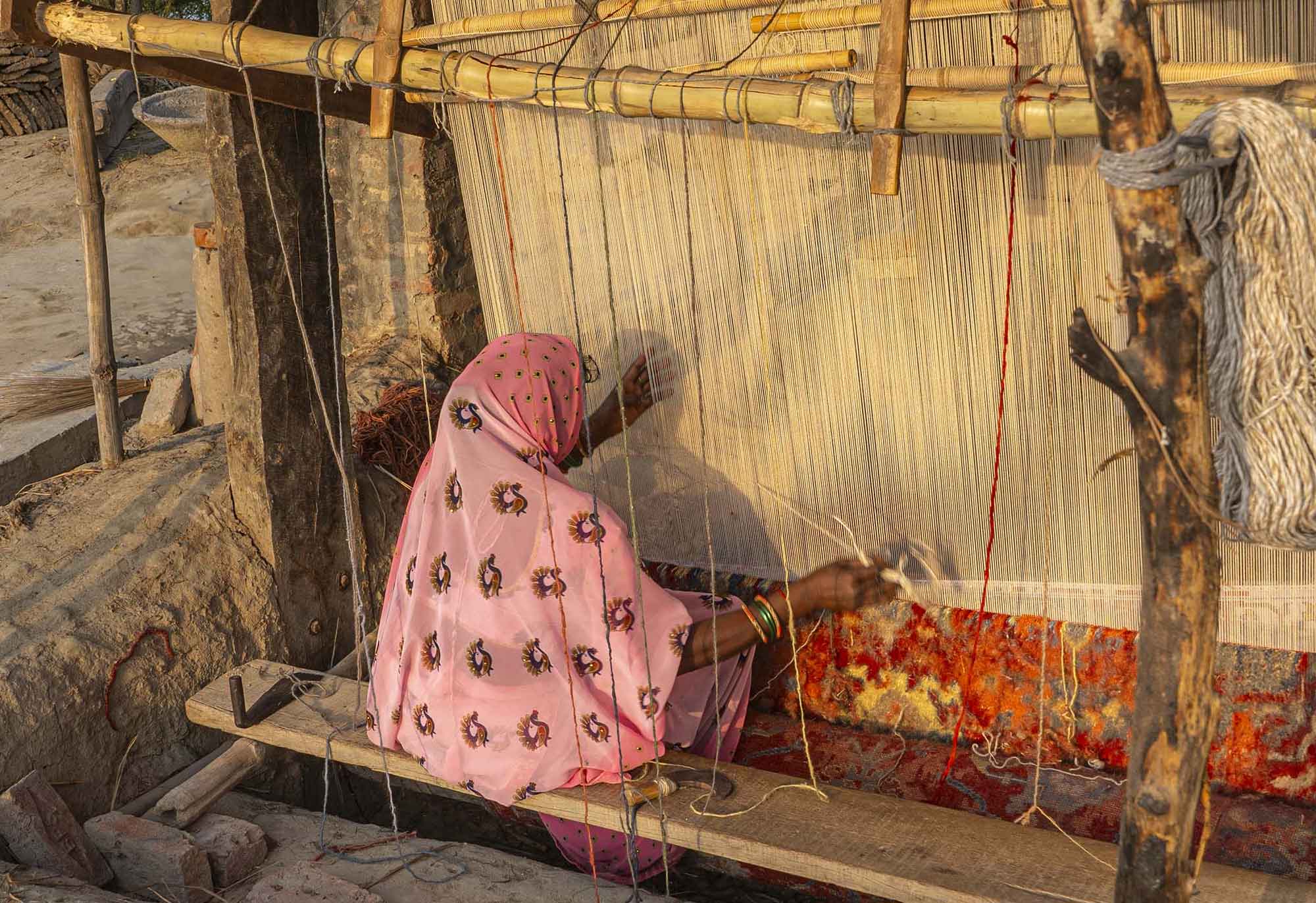 A woman in a pink patterned shawl weaves on a traditional loom, focusing intently. The scene conveys skill and dedication in a rustic setting.