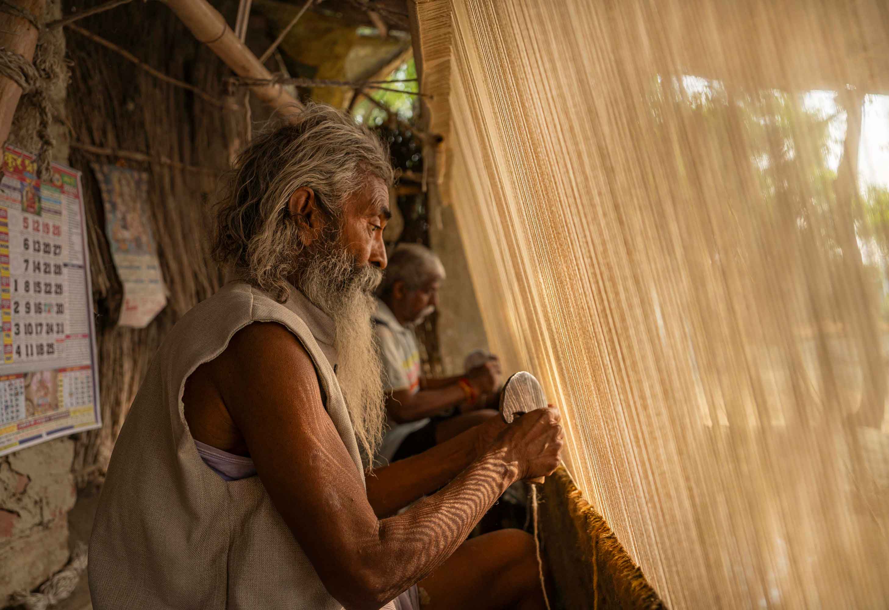 Elderly man weaving fabric by hand on a wooden loom, bathed in warm sunlight. A calendar hangs on a rustic wall, adding to the serene atmosphere.