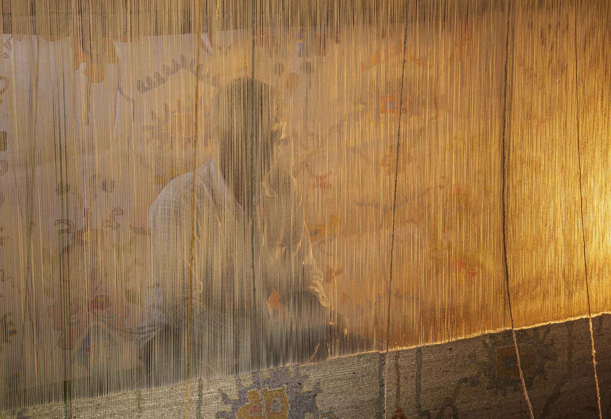 A person is seen through vertical threads of a loom, with warm lighting casting a serene glow. The background features a colorful, abstract pattern.