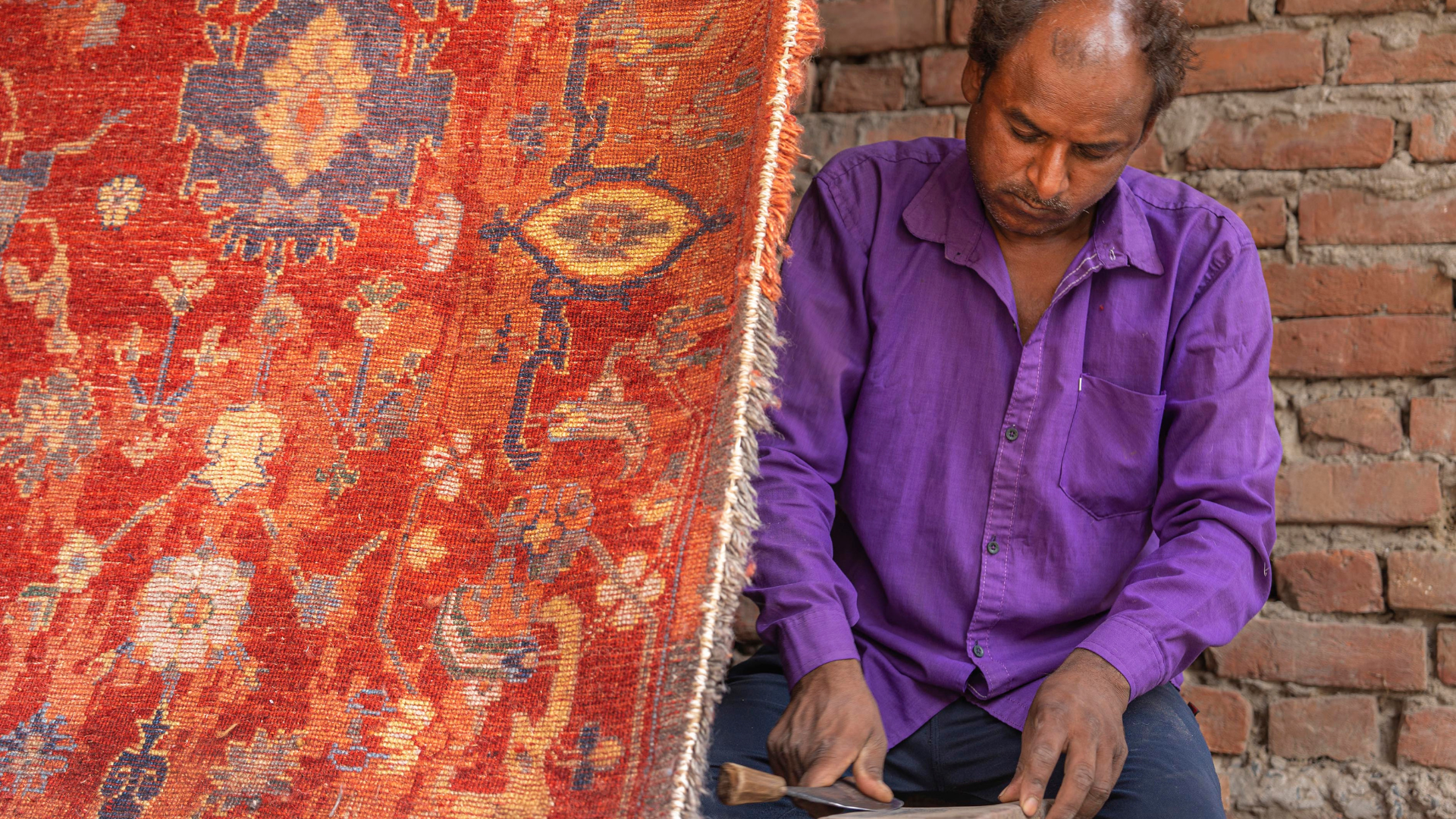 A man in a vibrant purple shirt concentrates on crafting a tool in front of a colorful, intricately patterned red carpet, against a brick wall backdrop.