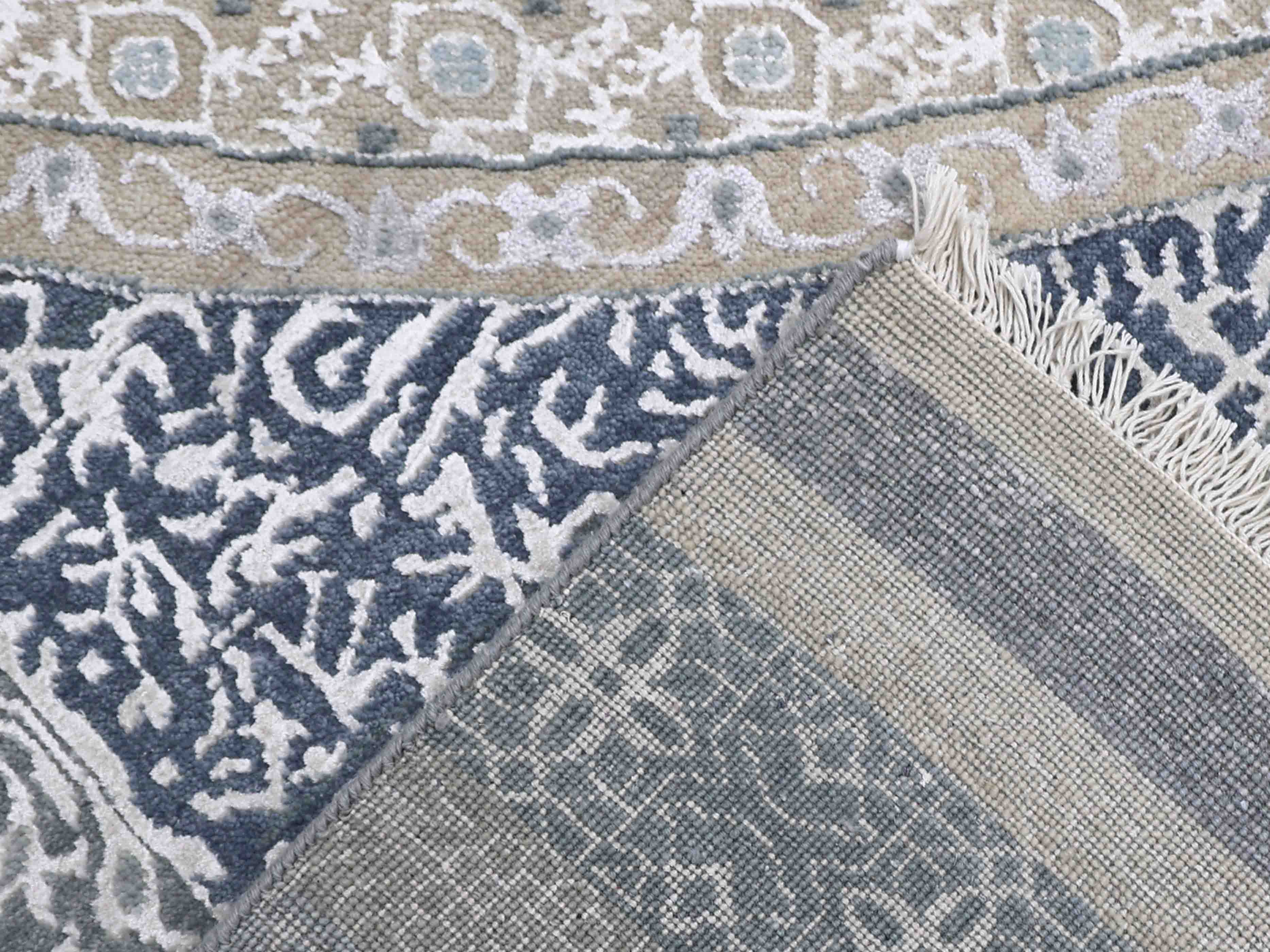 Close-up of intricately designed rug with blue and beige floral patterns. The corner is folded to reveal the fringed underside, adding texture.