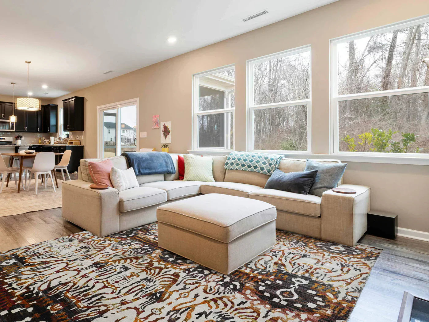 Bright living room with a large beige sectional sofa adorned with colorful pillows. A patterned rug adds warmth. Open kitchen and dining area in the background.