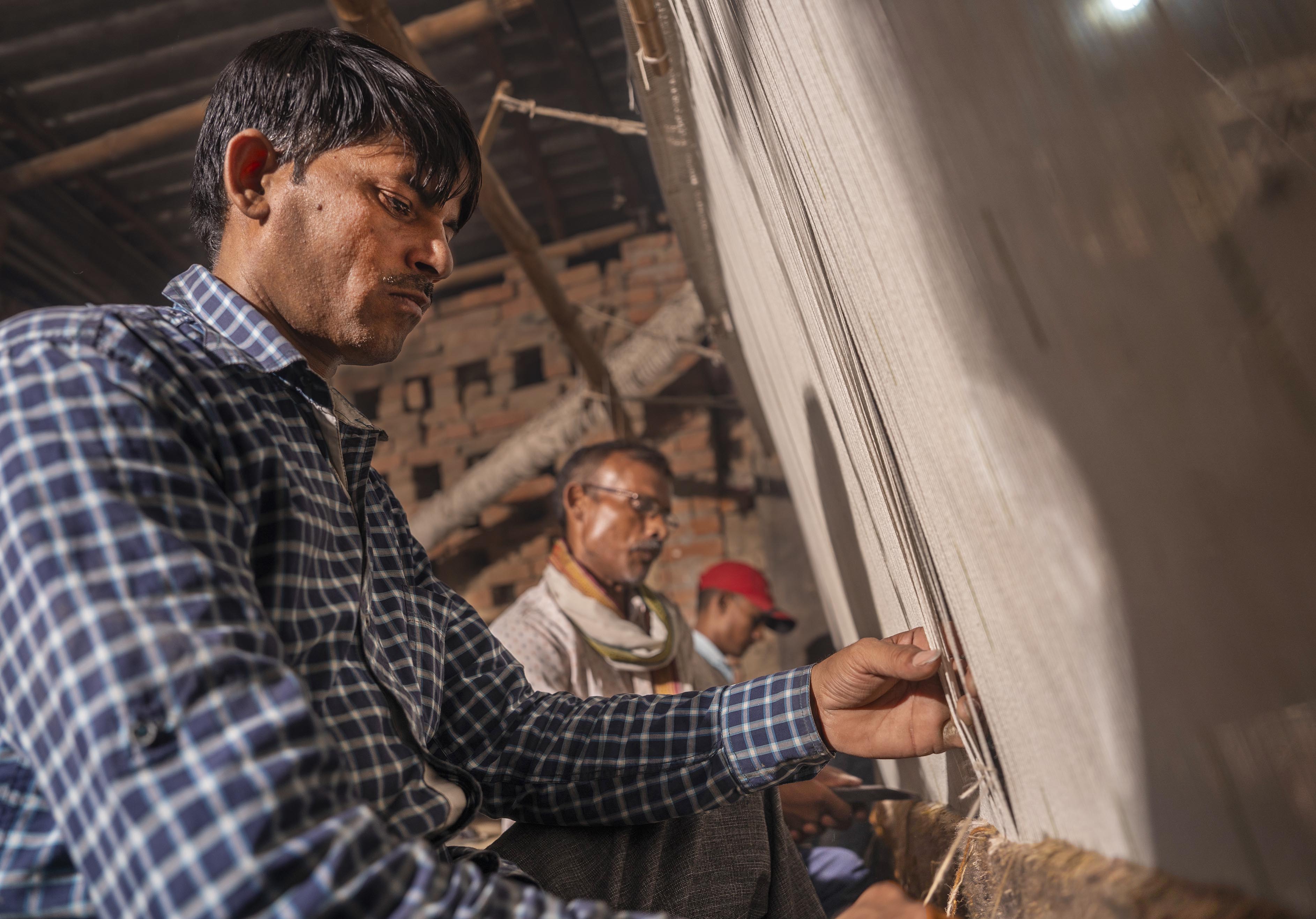 Man focused on weaving at a loom in a rustic workshop, with two men in the background. The setting is warm and conveys concentration and craftsmanship.