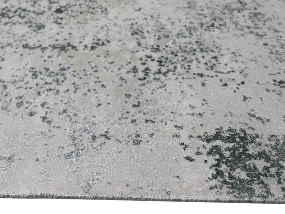 Close-up of a textured rug with an abstract design. The surface features scattered dark green speckles on a light gray background, giving an earthy, artistic feel.