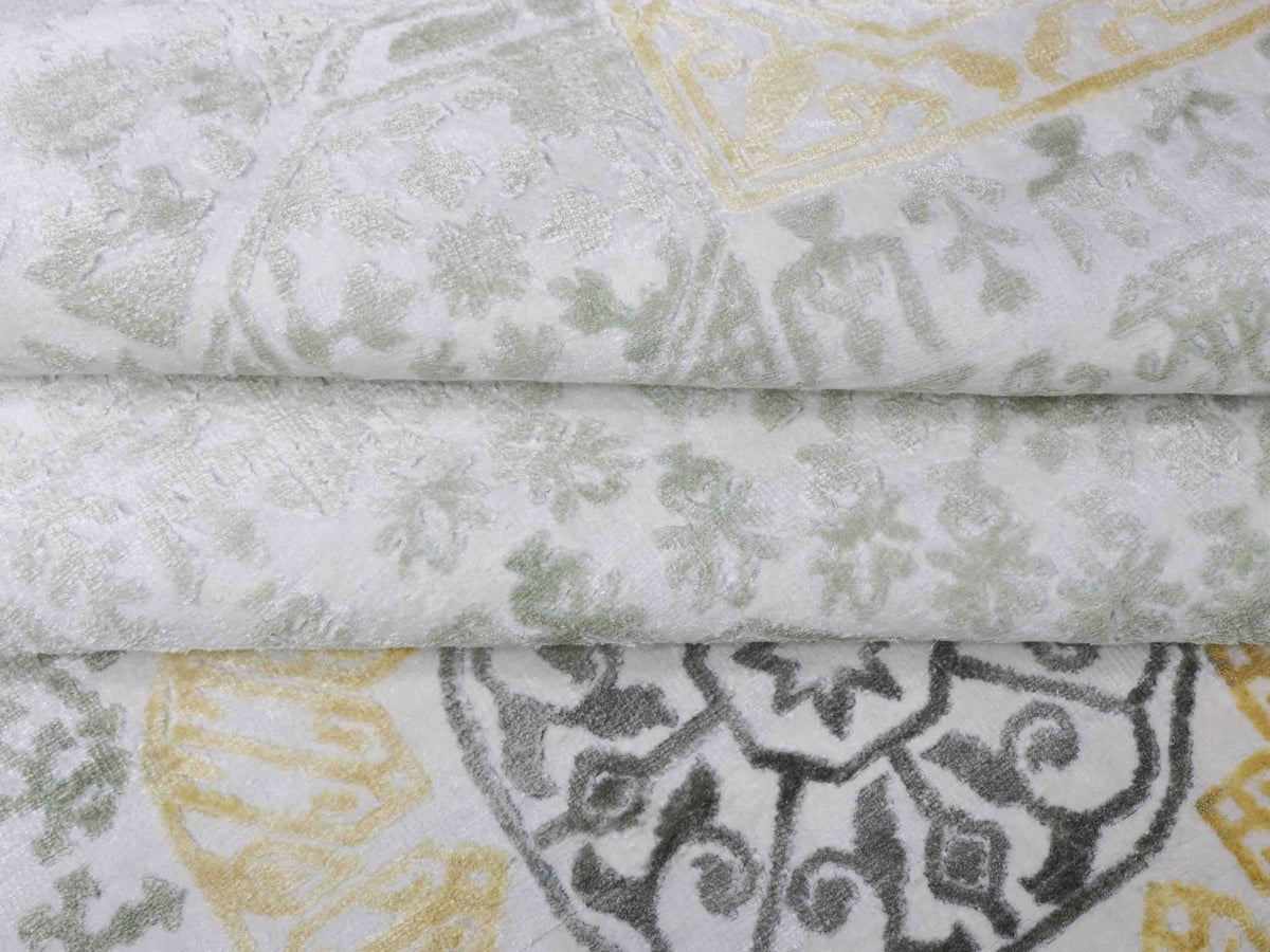 Close-up of folded fabric with intricate patterns in muted green, yellow, and gray tones. The design includes floral and geometric motifs, conveying elegance.