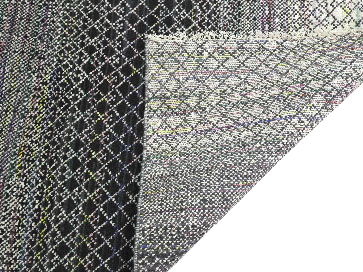 Close-up of a textured, multicolored woven rug with a diamond pattern. The fringed edge is folded over, revealing a similar inverse design.